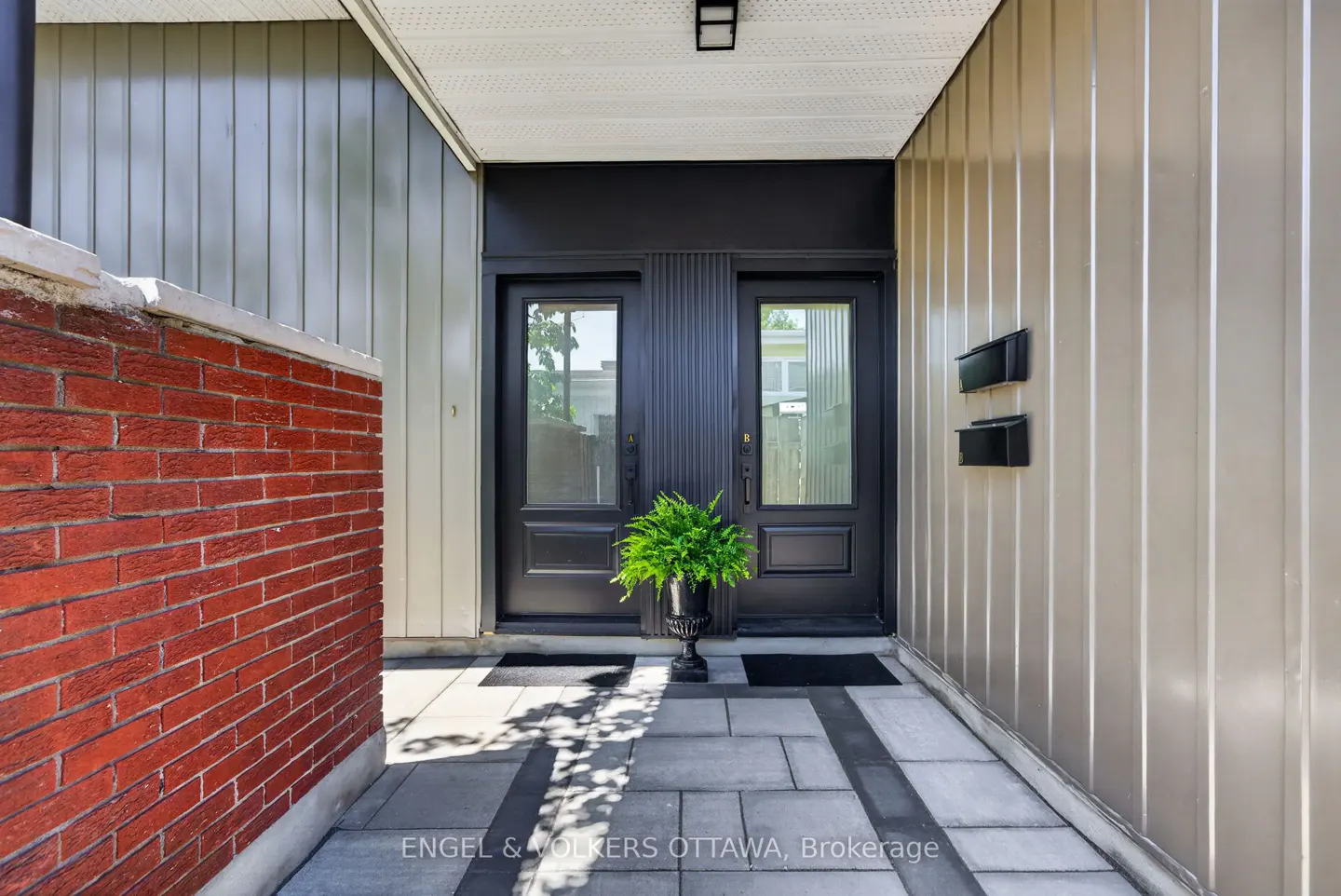Modern entryway with two black doors, a potted fern, red brick wall, and beige vertical siding on a stone patio.