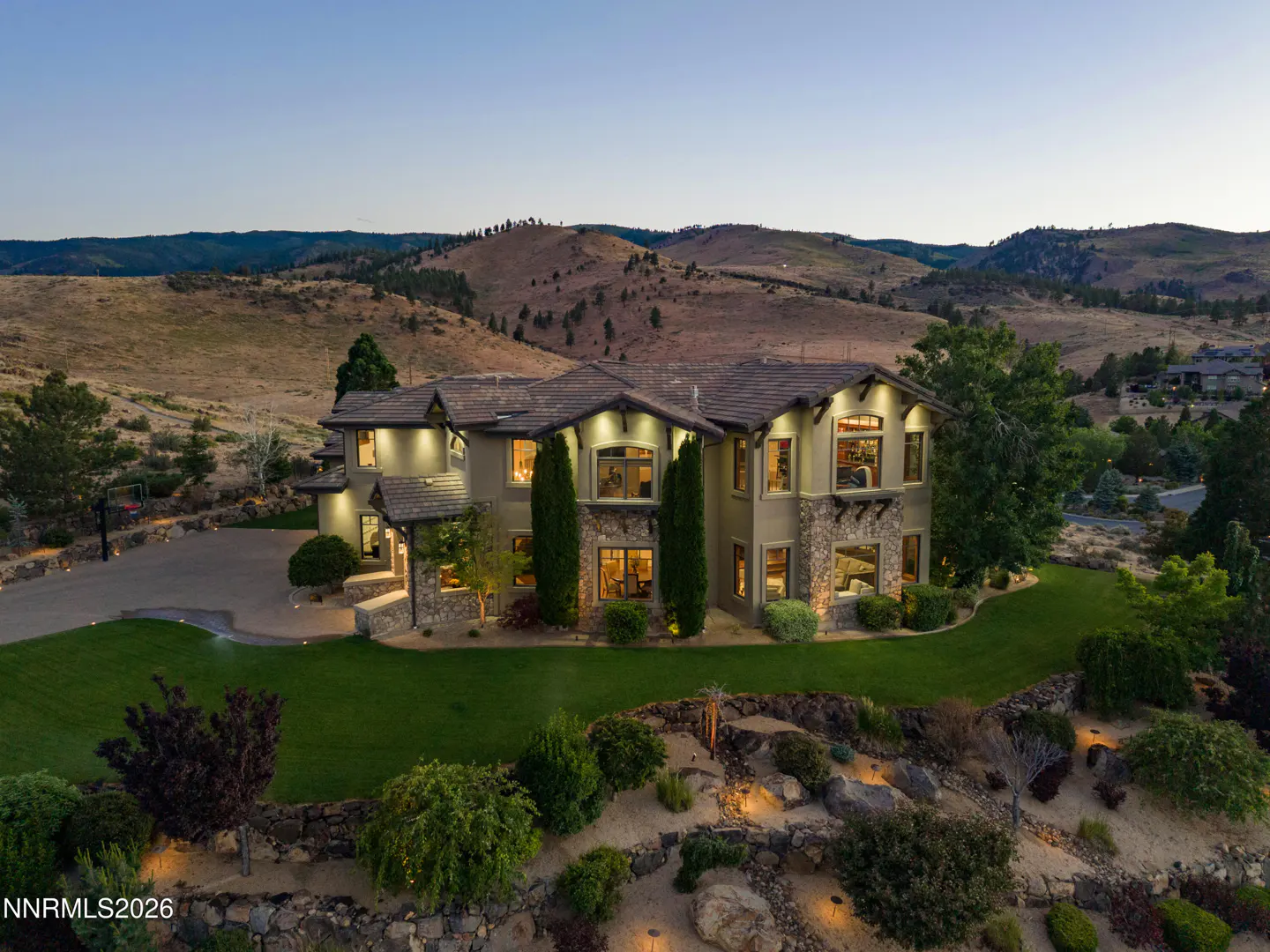 Aerial view of a large, multi-story stucco and stone home at dusk, set against rolling hills with a landscaped front yard.