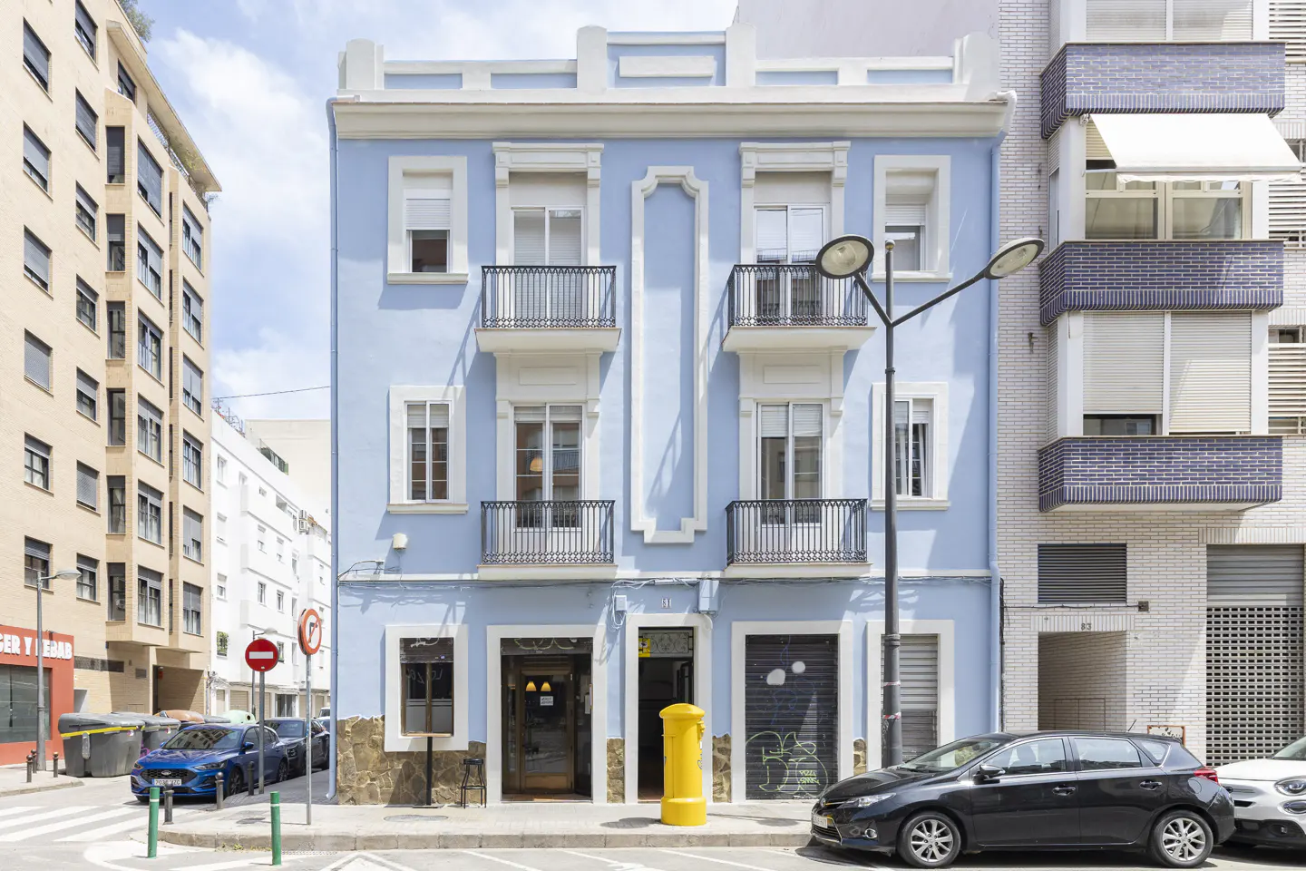Three-story light blue building with white trim and black iron balconies. A yellow post box sits on the sidewalk. Cars are parked on the street.
