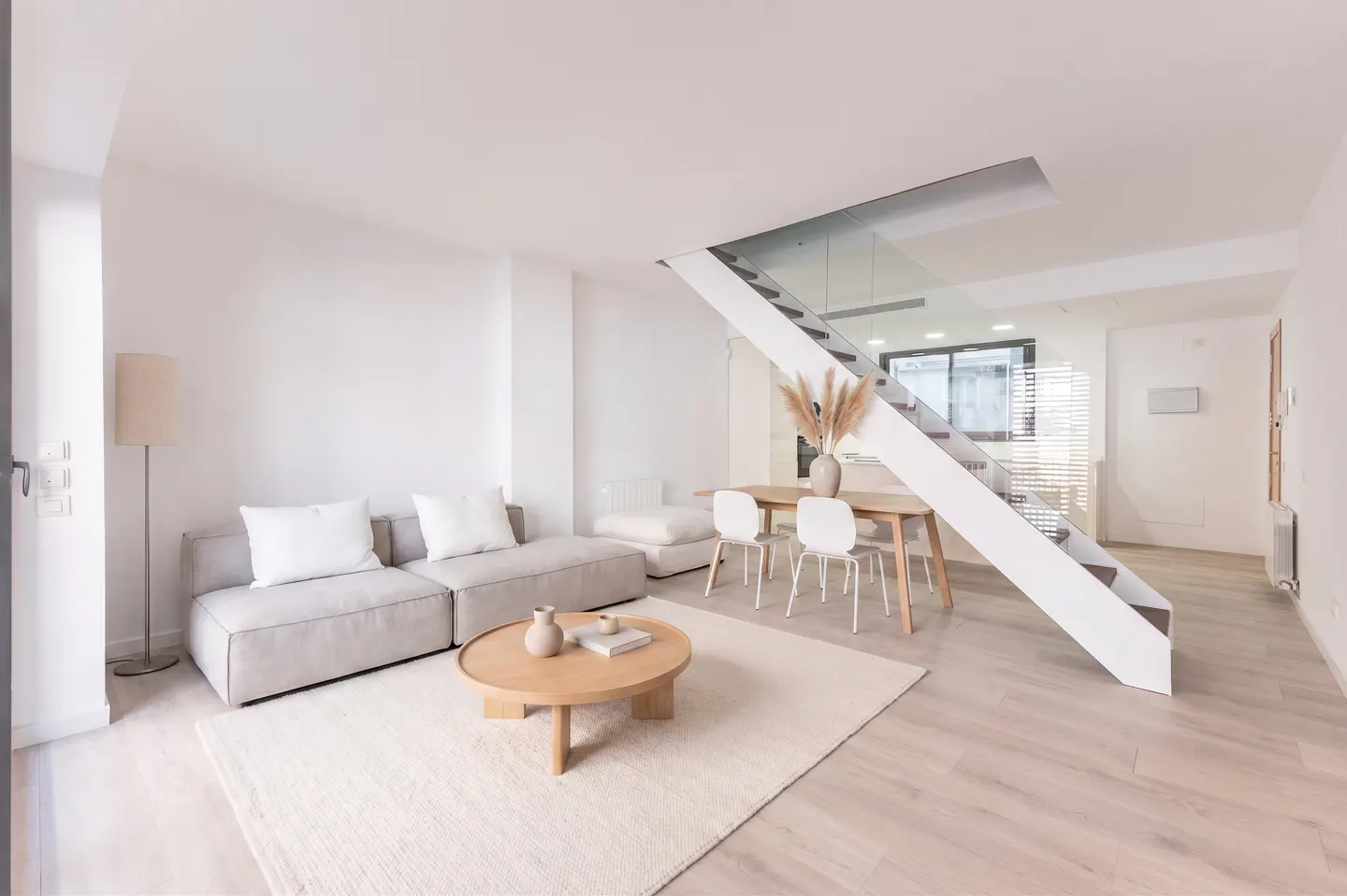 Bright, modern living room with a gray sofa, round wood table, and white stairs with glass railing. A dining table with chairs is visible.
