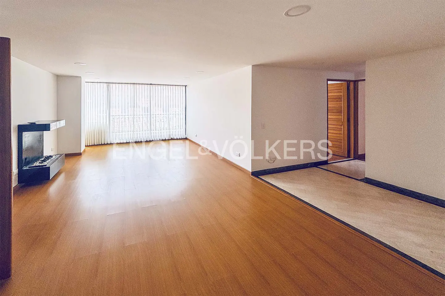 Bright, empty living room with wood floors, white walls, and a modern fireplace. Vertical blinds cover a large window. A doorway leads to another room.