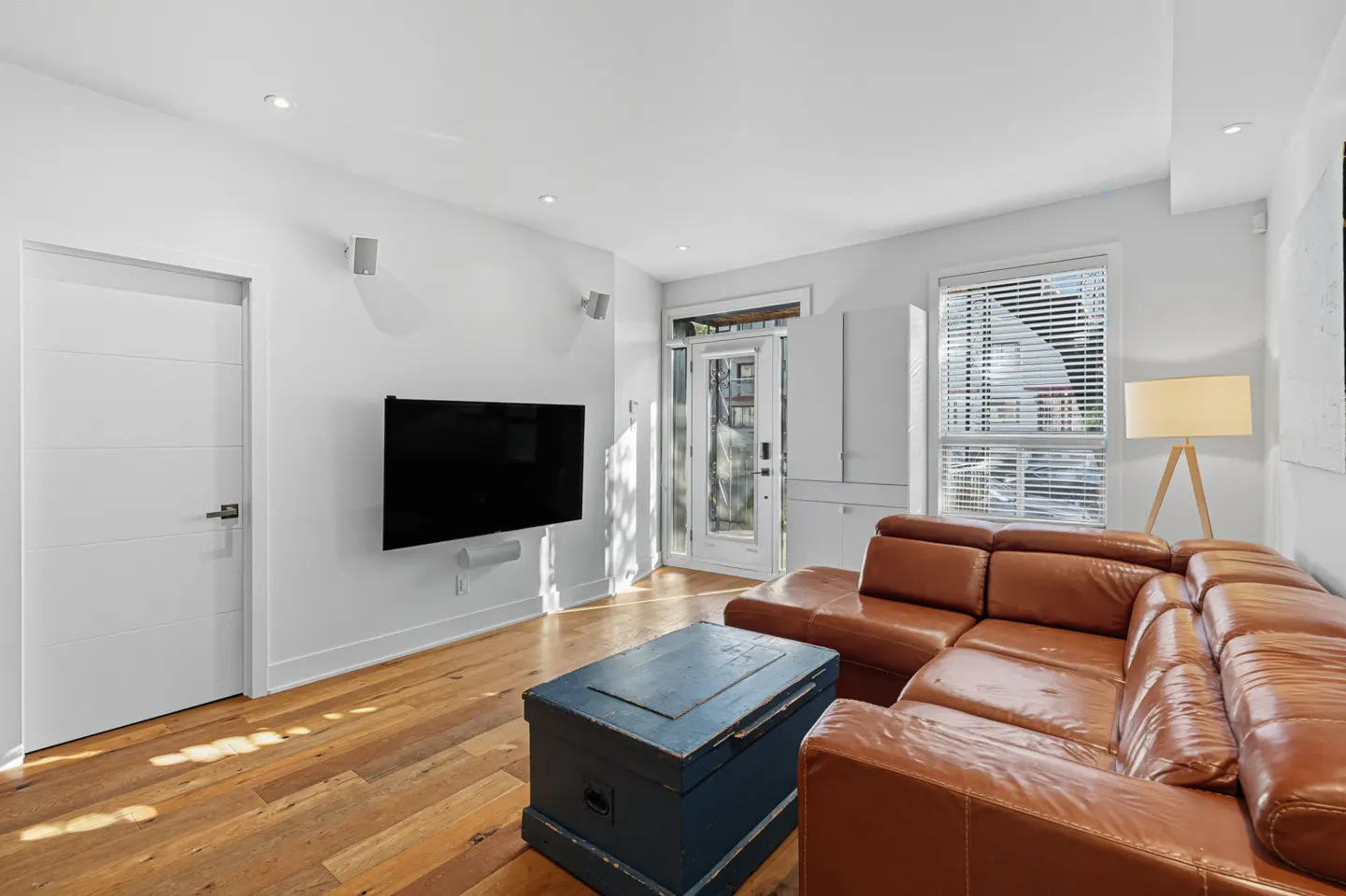 Bright living room with hardwood floors, a brown leather sectional, and a blue trunk coffee table. A TV hangs on the white wall.