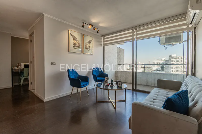 Bright living room with blue chairs, grey sofa, and city view through large windows. Artwork and a coffee table add to the modern decor.