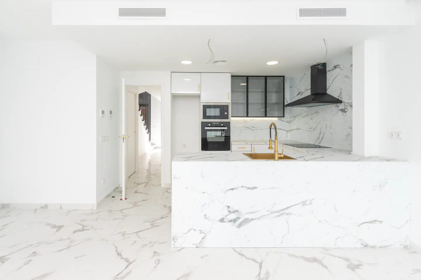 Bright, modern kitchen with white marble counters and floors. Black cabinets and range hood accent the white walls. A gold faucet adds a touch of luxury.