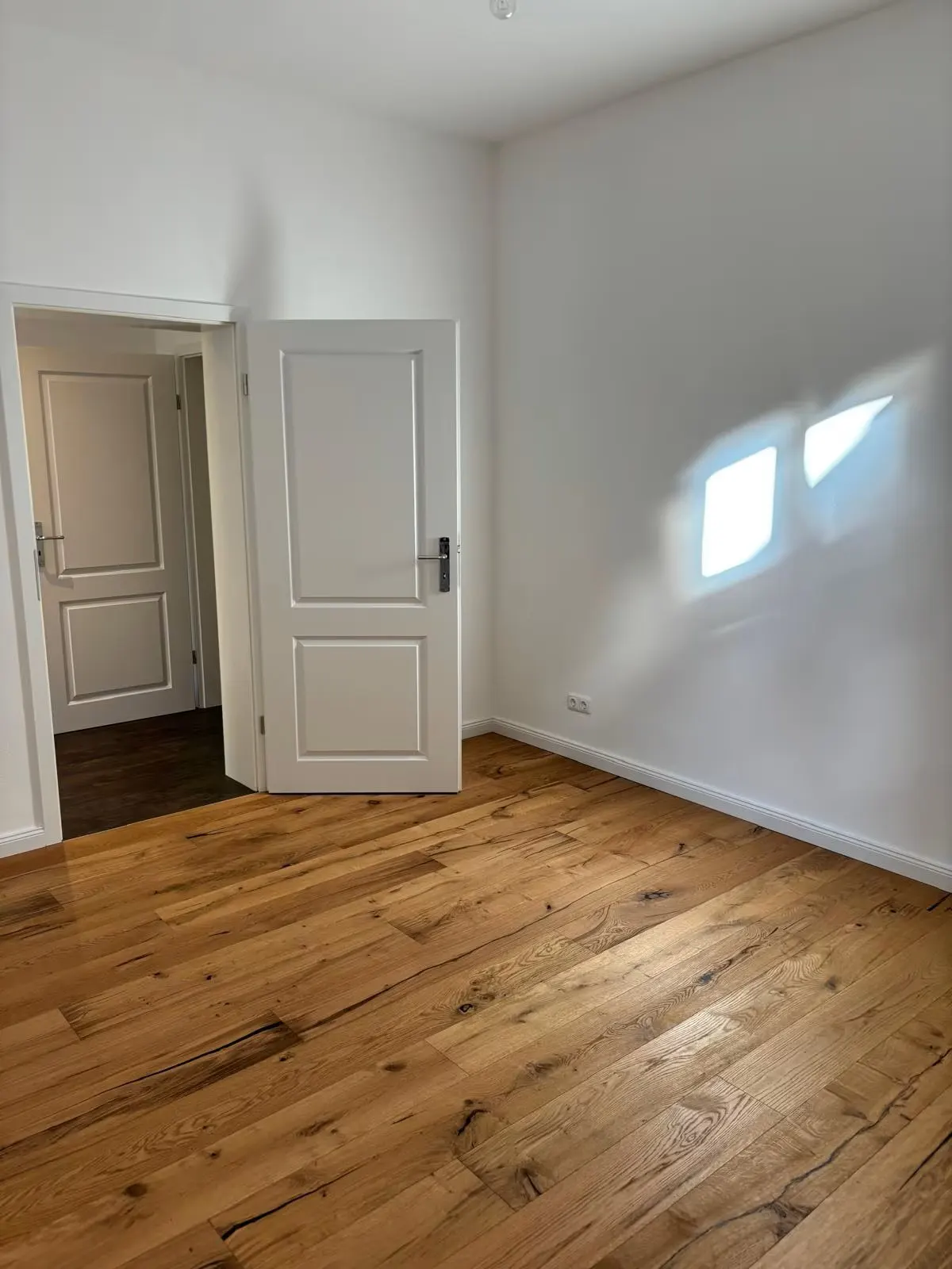 Bright, empty room with hardwood floors, white walls, and two white doors. Sunlight shines through a window.