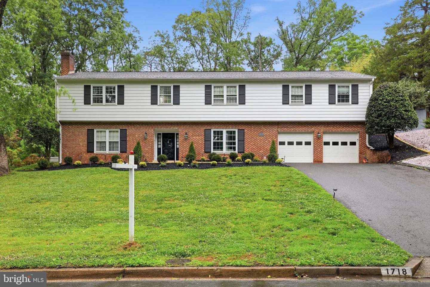 Two-story house with red brick and white siding, black shutters, and a green lawn. Two-car garage and black driveway.