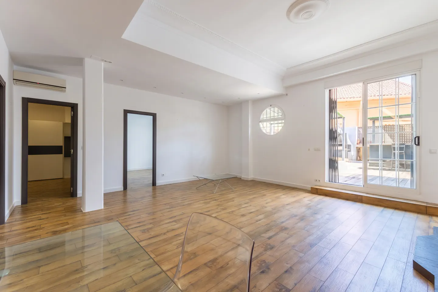 Bright, empty room with wood floors, white walls, and two doorways. A glass table and sliding glass doors to a patio are visible.