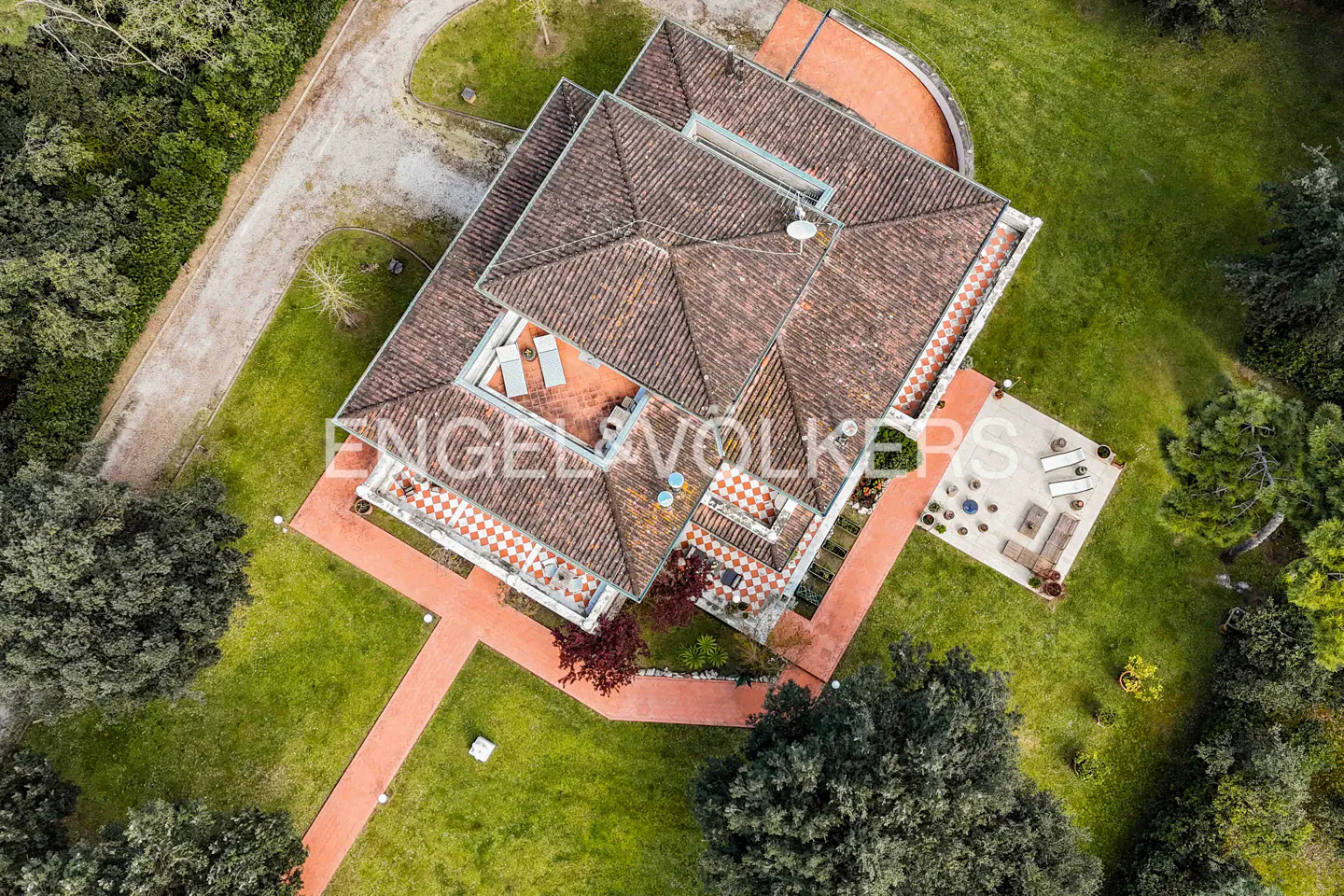 Aerial view of a villa with a brown tile roof, red brick paths, and green lawn surrounded by trees. Patio furniture is visible.