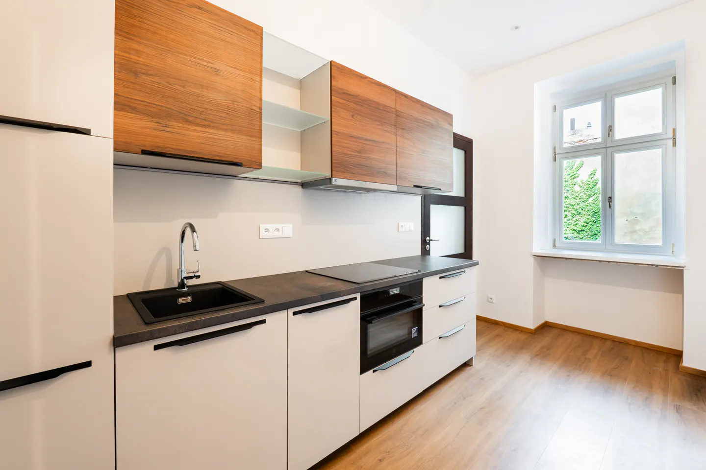 Modern kitchen with white lower cabinets, wood-grain upper cabinets, black countertops, and a window with a view of greenery.