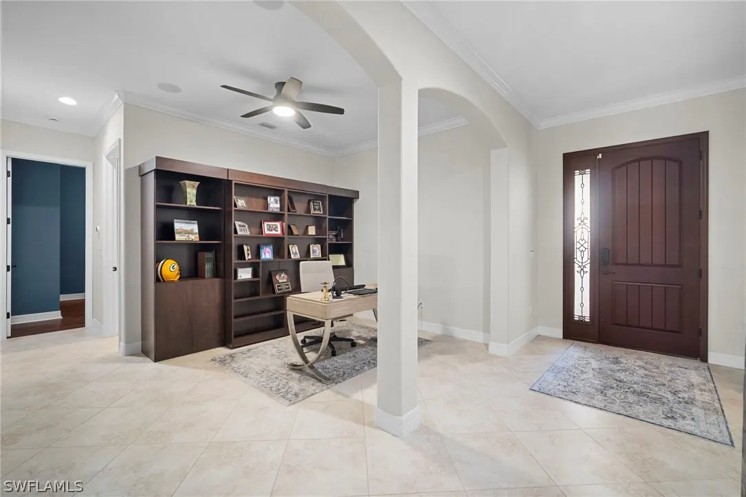 Bright home office with a large brown bookcase, desk, and chair. A brown front door with glass is on the right. Beige tile floor.