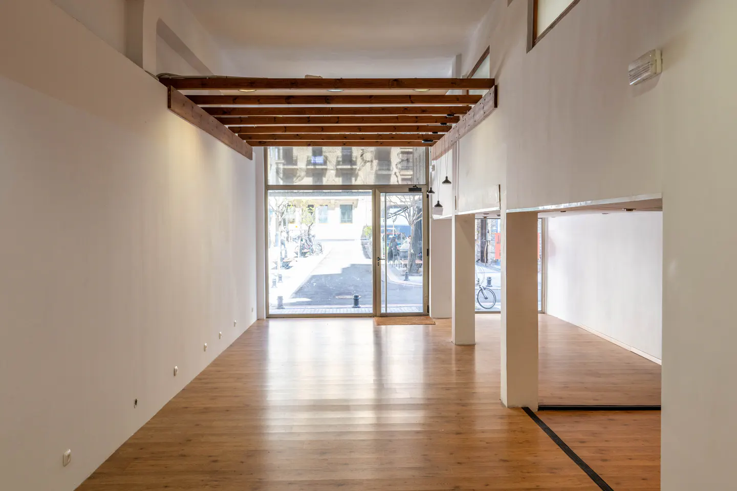 Bright, empty commercial space with hardwood floors, white walls, and a large glass storefront. Wooden beams accent the high ceiling.