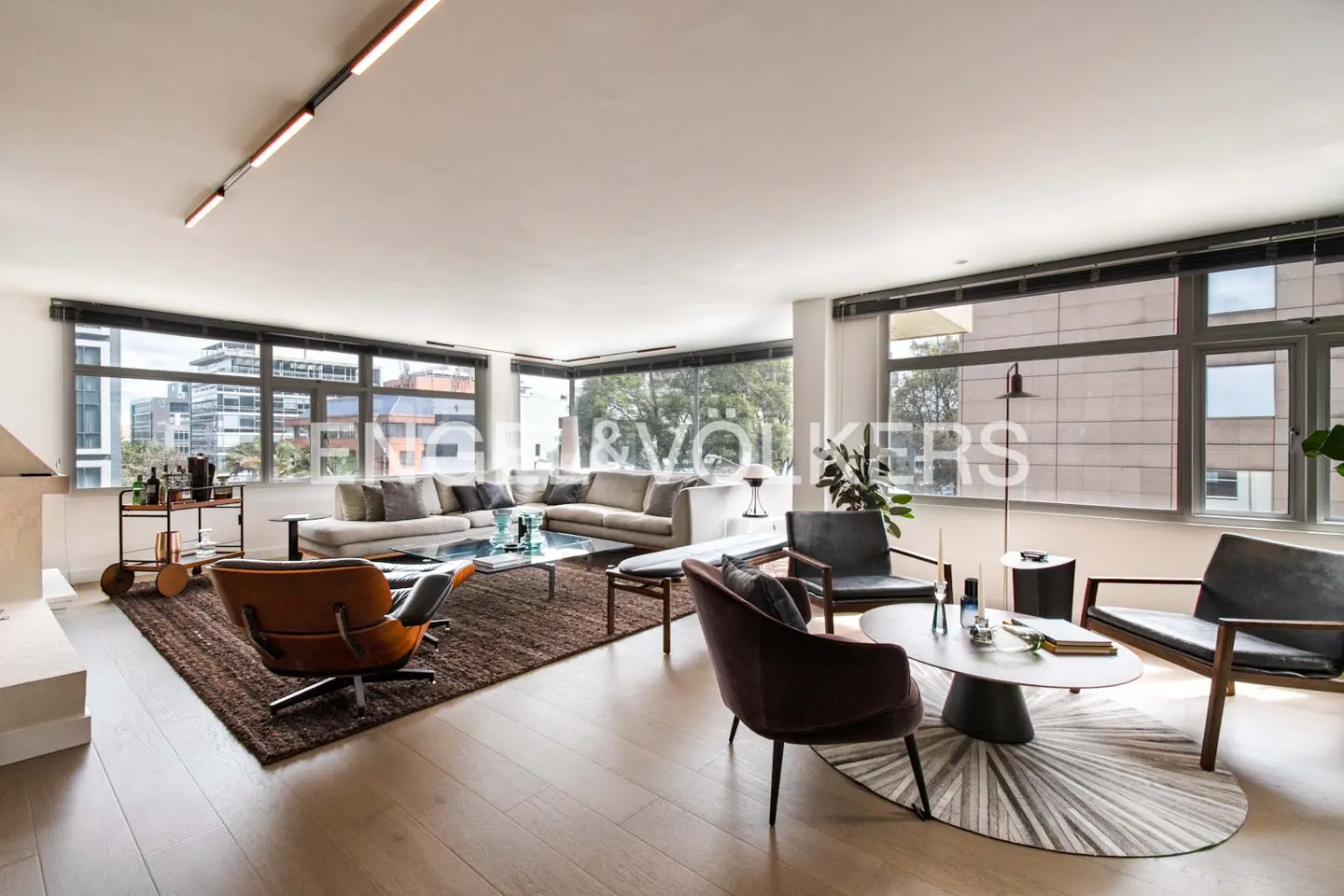 Bright, modern living room with large windows, a gray sectional sofa, and a brown leather chair. A bar cart sits near a fireplace.