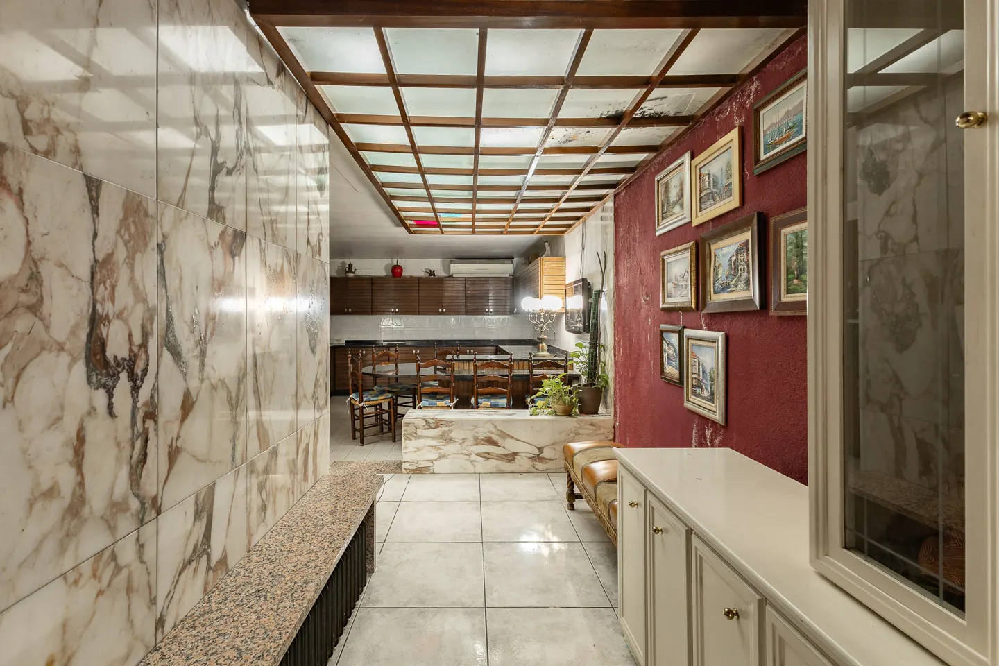 Hallway with marble walls, red accent wall with framed art, grid ceiling, and view into a dining area with wood cabinets.