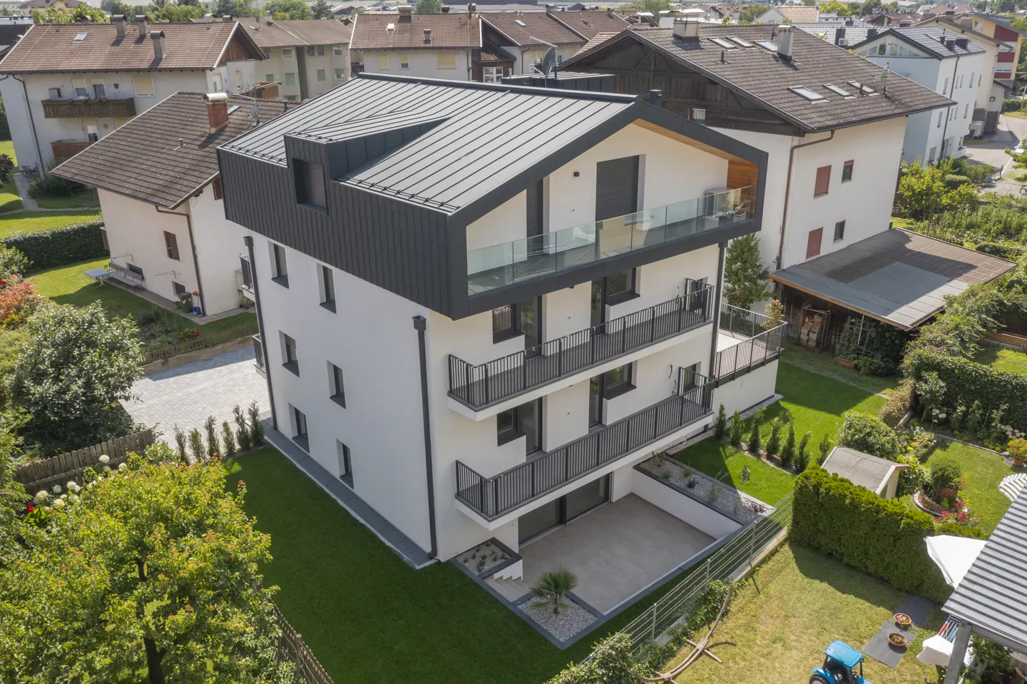Aerial view of a modern, three-story white building with a dark gray roof and black balconies, surrounded by green lawns and trees.