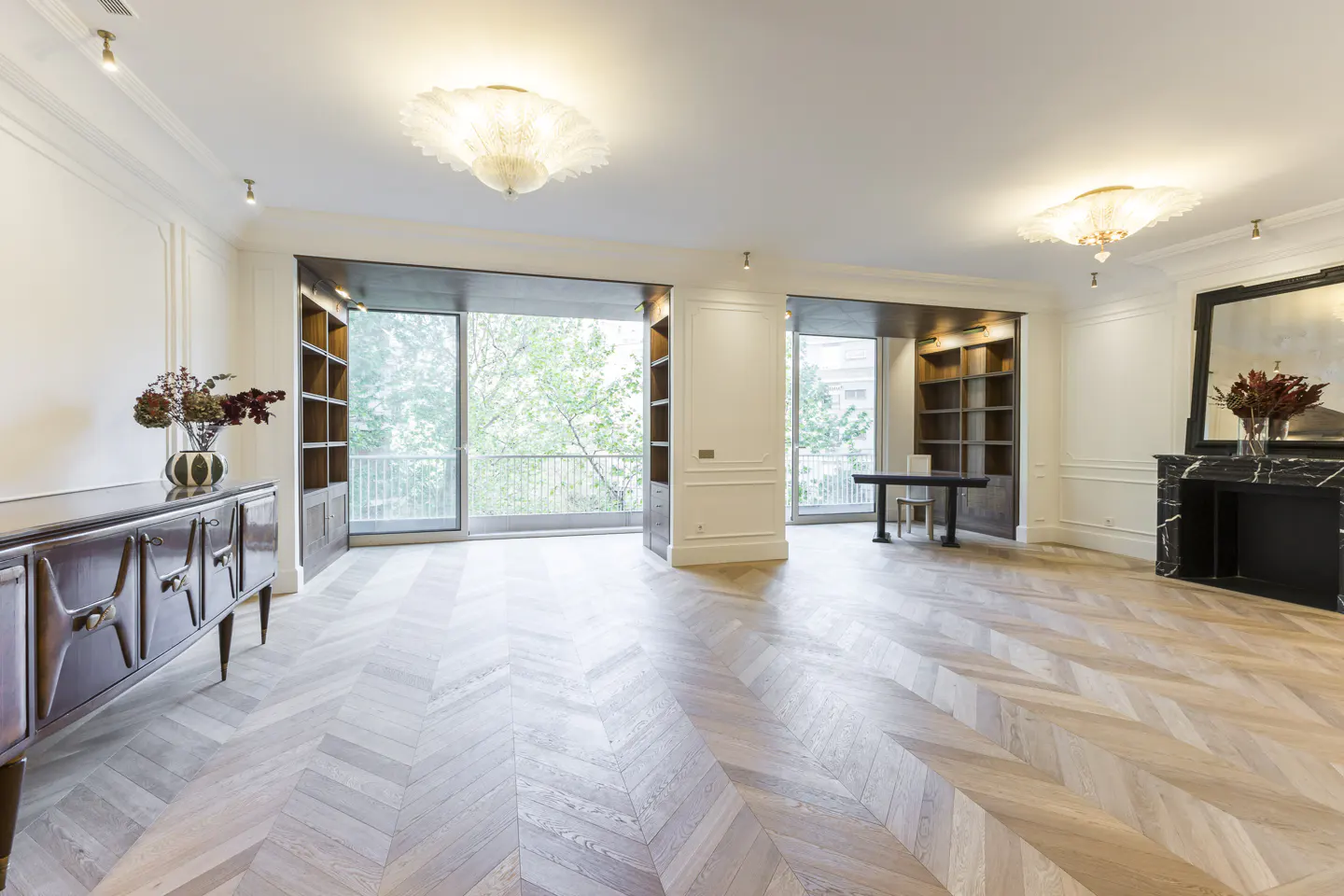 Bright, empty living room with herringbone wood floors, white walls, and large windows overlooking trees. A dark wood cabinet and fireplace are visible.