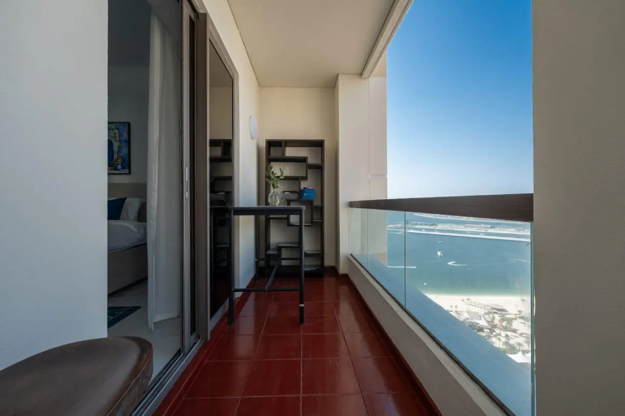 Balcony with red tile floor, glass railing, and ocean view. A black table and shelf are against the wall. Sliding glass doors lead to a bedroom.