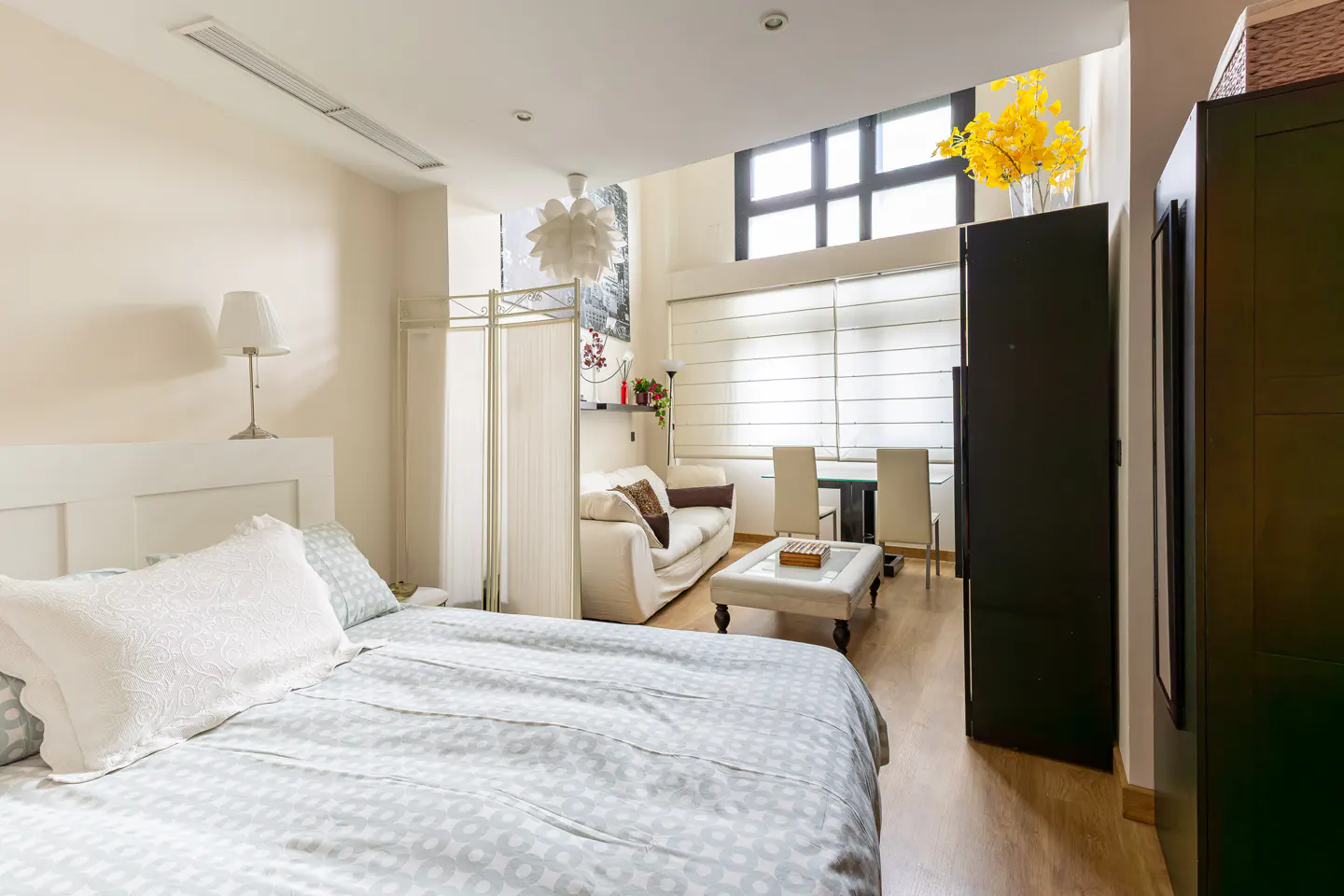Bright studio apartment with a bed, white sofa, dining table, and large windows. Yellow flowers add a pop of color.