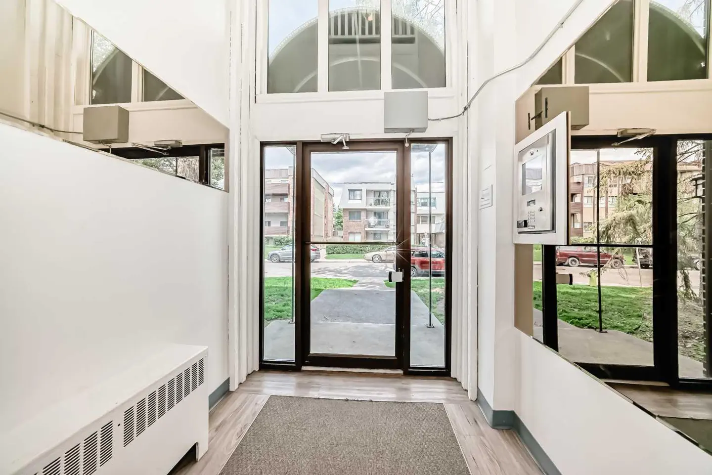 Apartment building entrance with glass doors, white walls, and a gray mat. Outside, there's a street with cars and buildings.