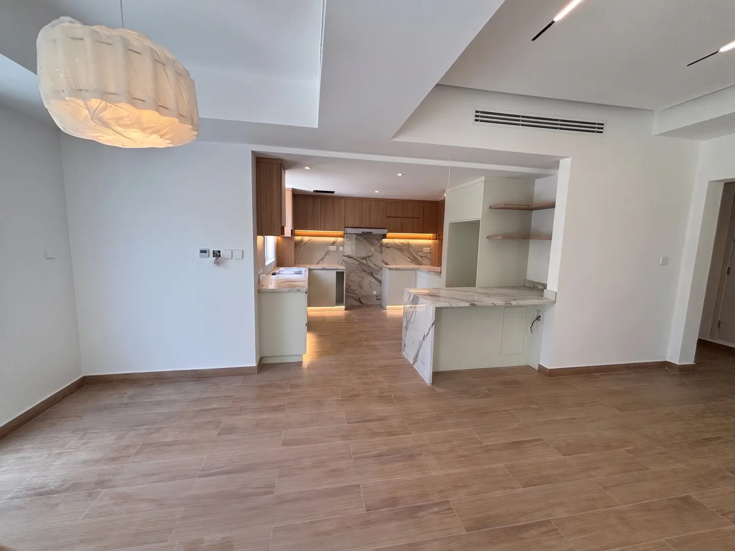 Open-concept kitchen and living room with wood-look tile flooring. White walls, marble countertops, and wood cabinets. Modern lighting.