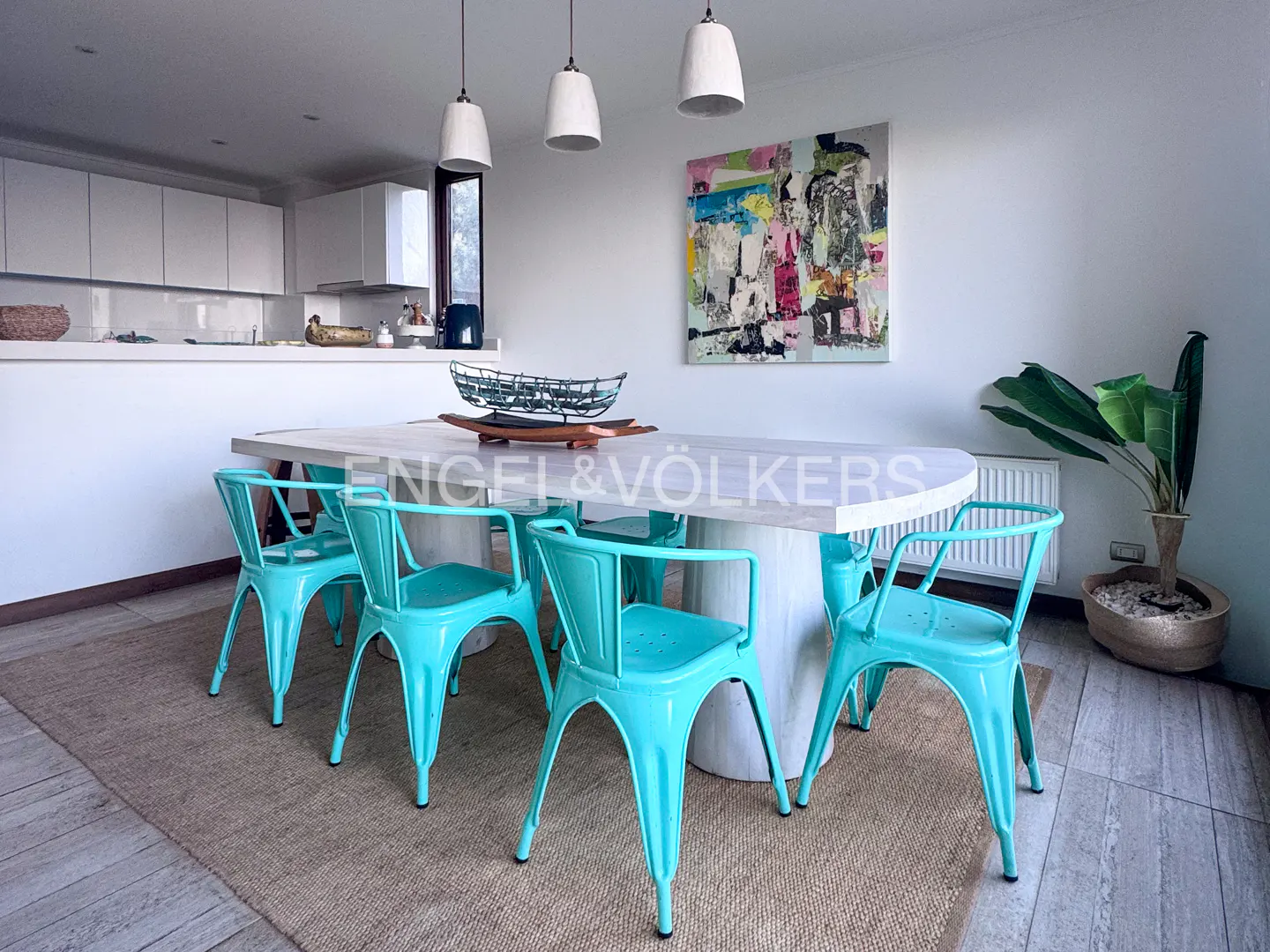 Bright dining area with turquoise chairs around a white table. Modern art hangs on the wall. Kitchen in the background.