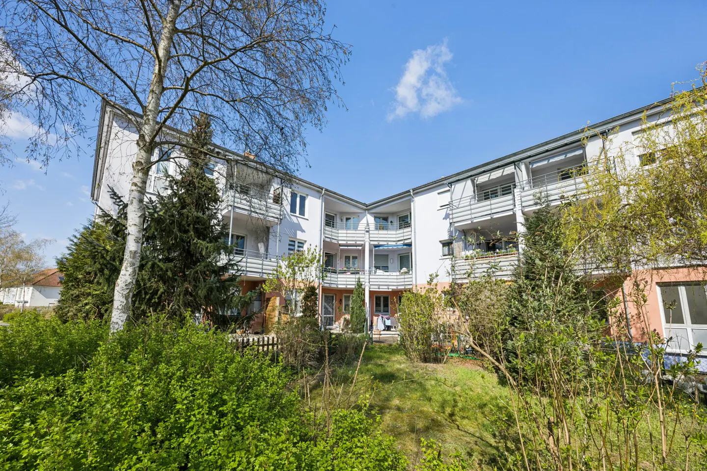 Exterior view of a white apartment building with balconies, surrounded by green trees and bushes under a blue sky.