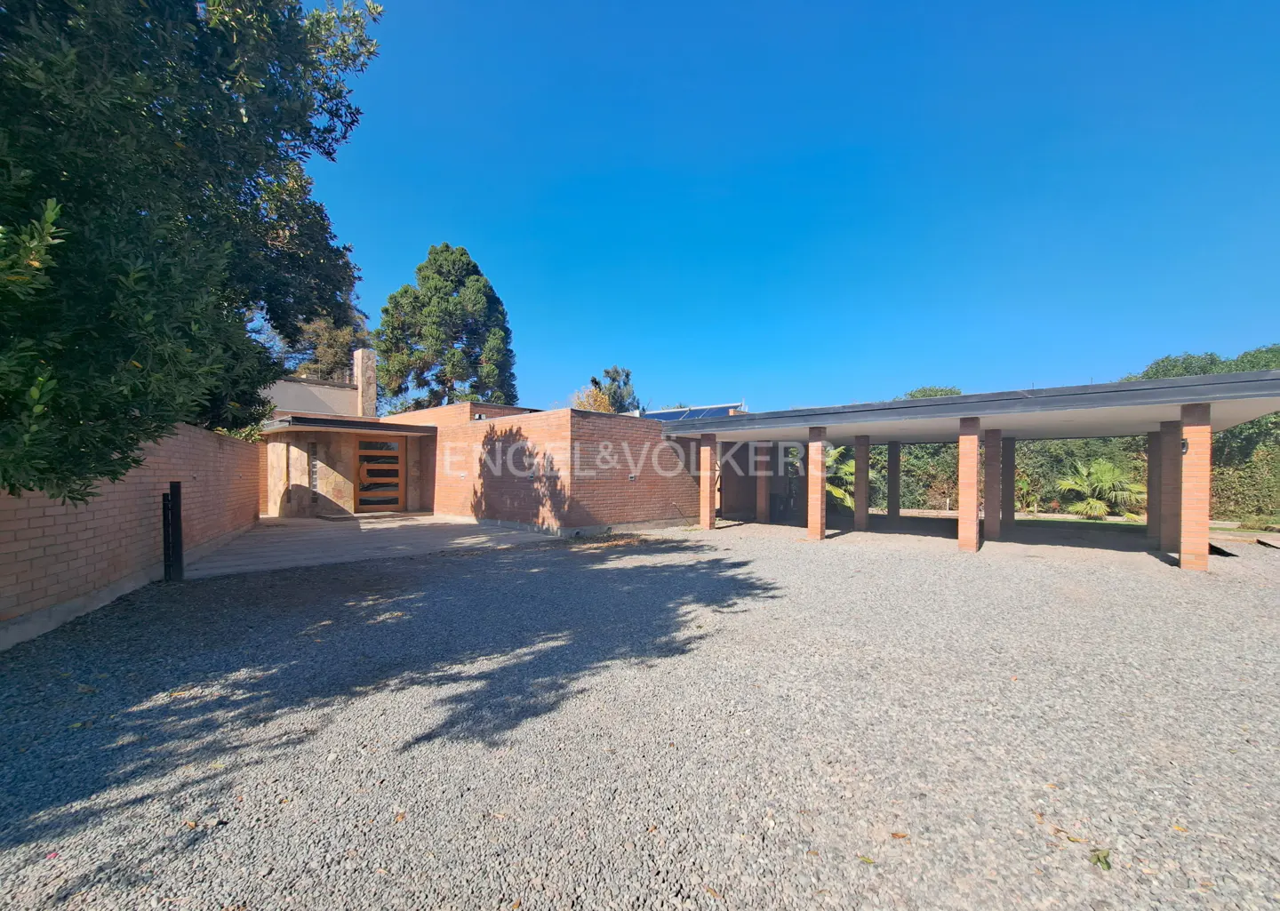 Exterior view of a brick house with a carport and gravel driveway under a clear blue sky.