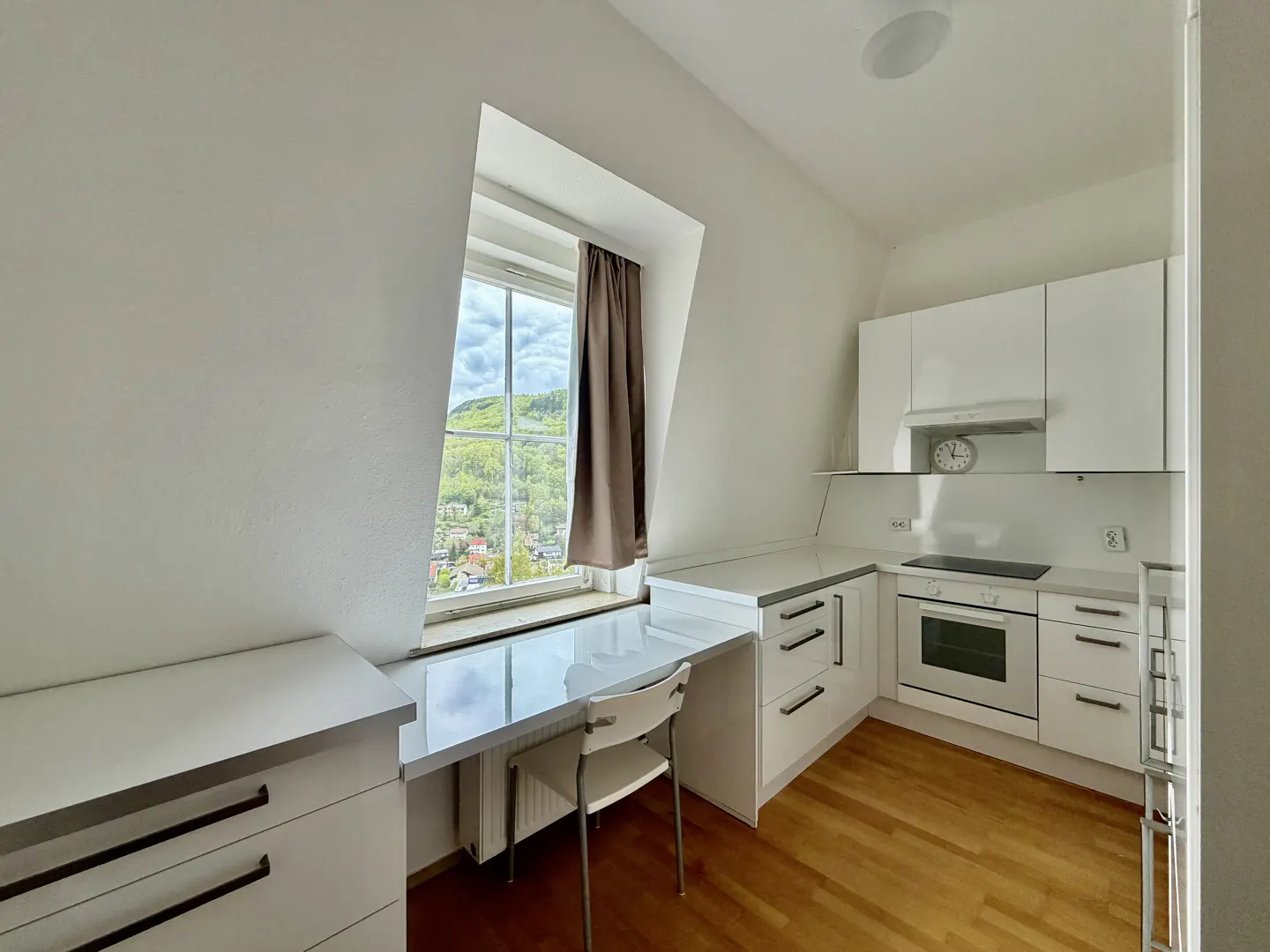 Bright, modern kitchen with white cabinets, countertops, and appliances. A large window with a brown curtain overlooks a green, hilly landscape.