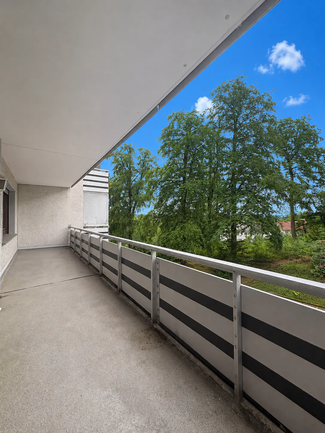Spacious covered balcony with a modern metal railing overlooking lush green trees under a bright blue sky.