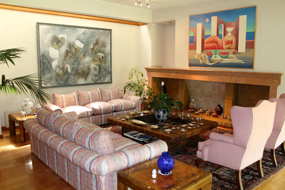 A living room with patterned sofas, a wooden coffee table, and a stone fireplace. Two paintings hang on the walls.