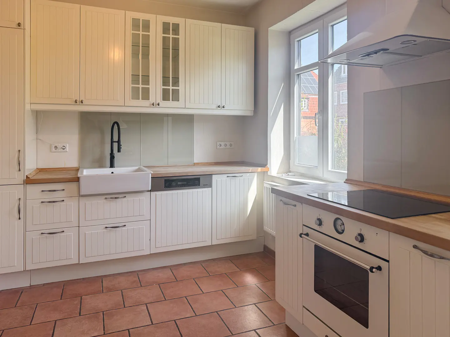 Bright kitchen with white cabinets, a farmhouse sink, and a black faucet. The floor is tiled in terracotta. A window lets in natural light.