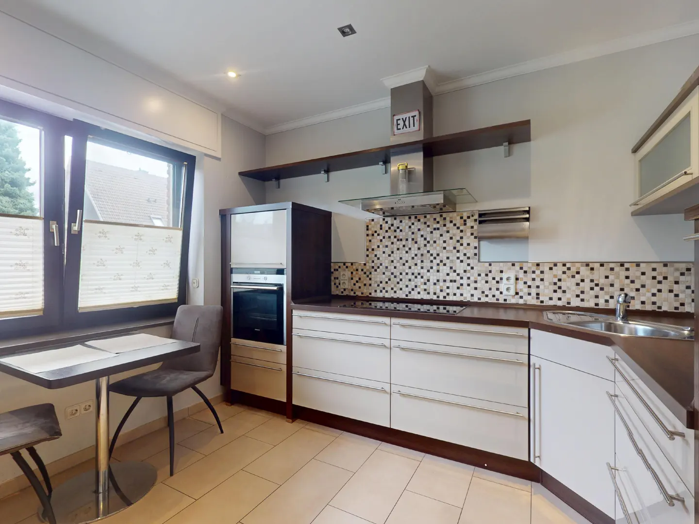 A modern kitchen with white cabinets, dark countertops, and a mosaic tile backsplash. A table and chairs sit near a window with blinds.