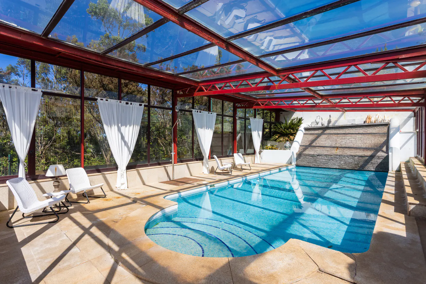 Indoor pool with glass ceiling and red beams. White curtains hang near windows overlooking trees. Lounge chairs sit poolside.