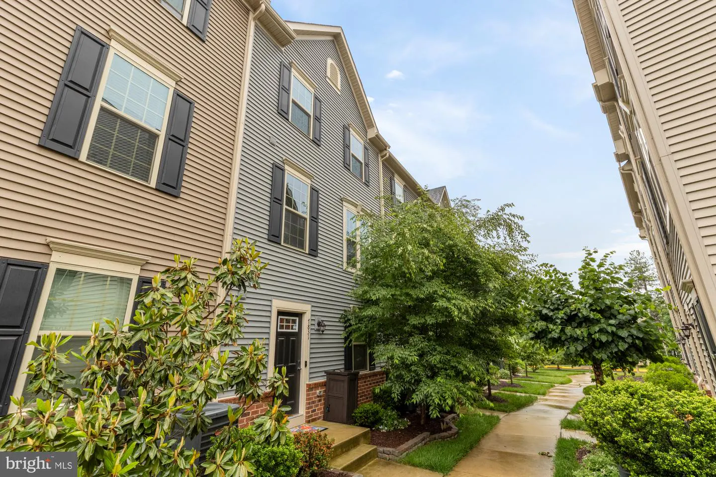 Exterior view of townhouses with tan and gray siding, black shutters, and green trees along a sidewalk.