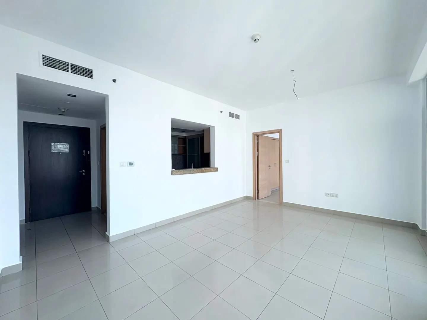 An empty room with white walls and gray tile floors, featuring a dark brown door and a kitchen pass-through.