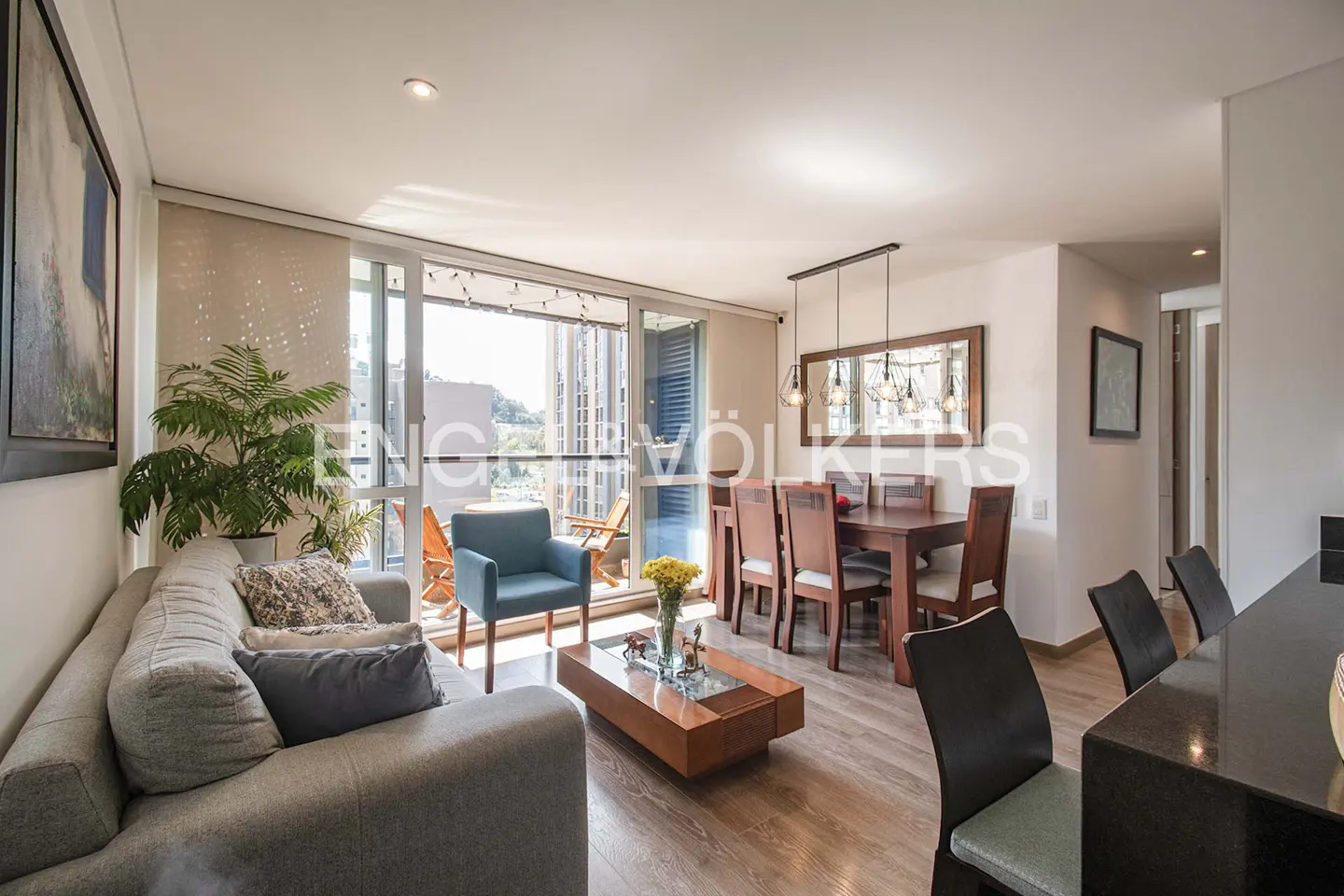 Bright, modern living room with a gray sofa, blue chair, wood table, and dining set. Balcony with city views.