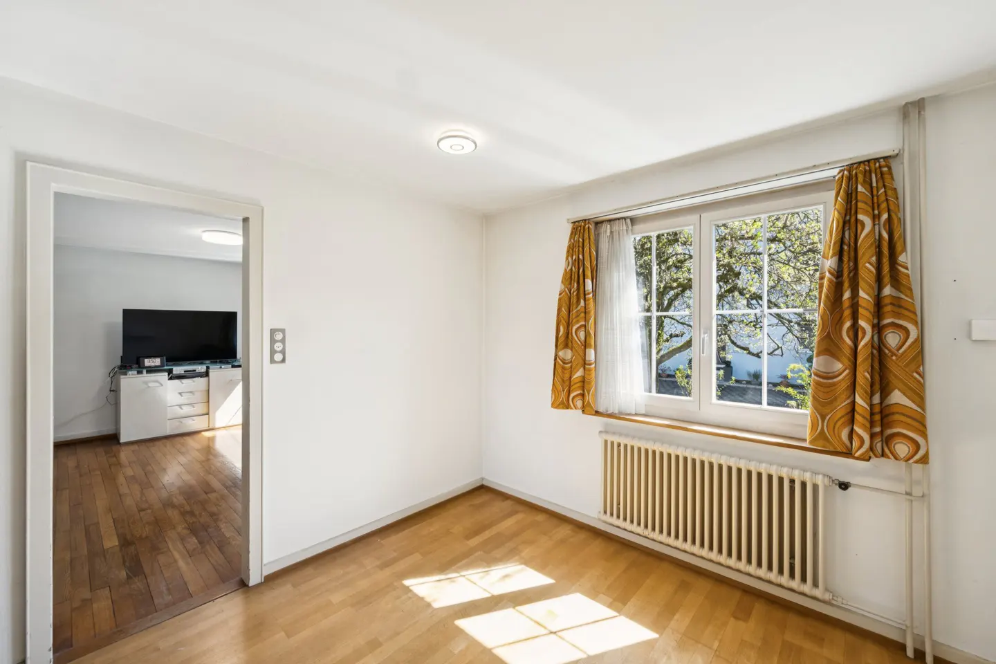 Bright room with wood floors, white walls, and a window with patterned curtains. A doorway leads to another room with a TV.