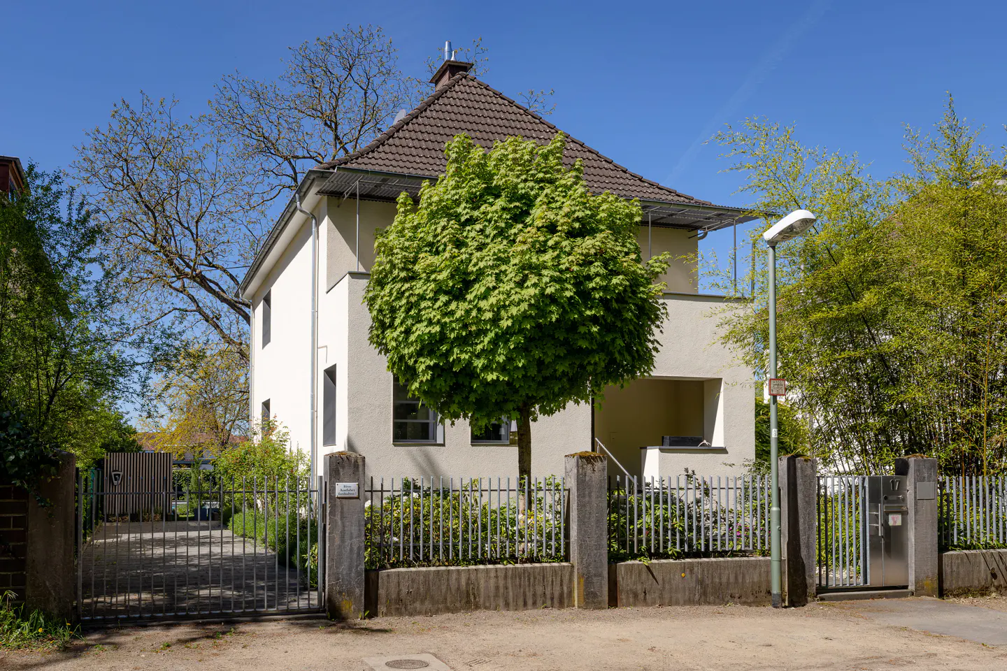Two-story white house with a brown roof, green tree in front, and a gray metal fence.