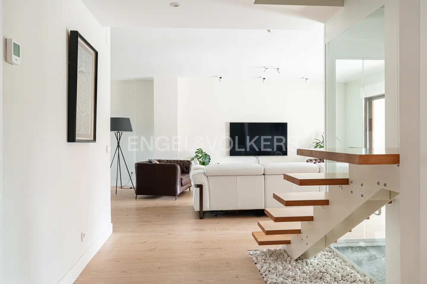 Bright, modern living room with white walls, light wood floors, and floating wood stairs. A white sofa and brown leather chair are visible.