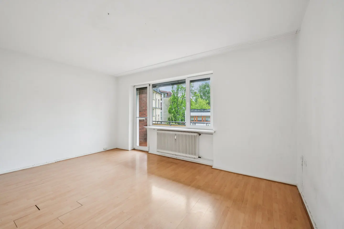 Bright, empty room with light wood floors, white walls, and a window overlooking greenery. A radiator sits beneath the window.