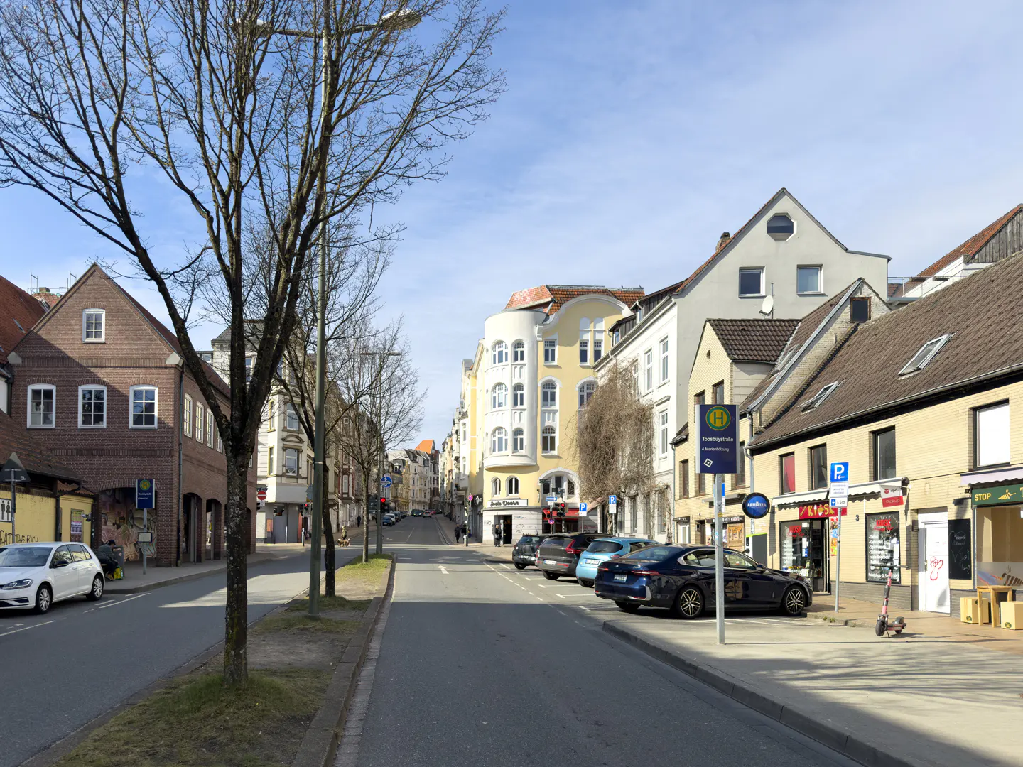 Street view of a European town with parked cars, shops, and brick buildings under a blue sky.