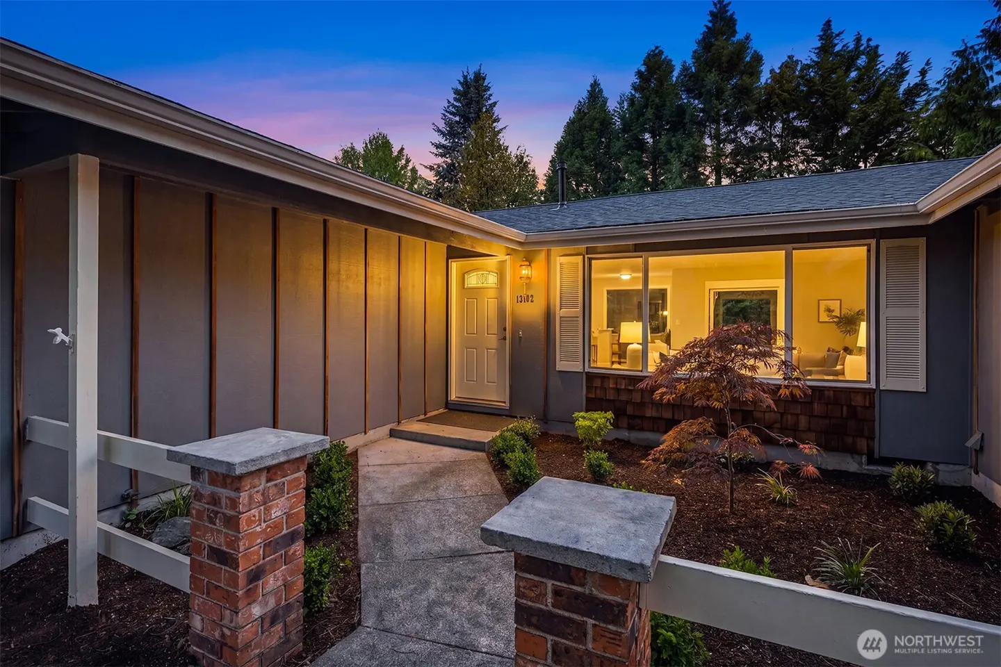 Exterior view of a gray house with a white front door, large window, and brick accents at dusk.