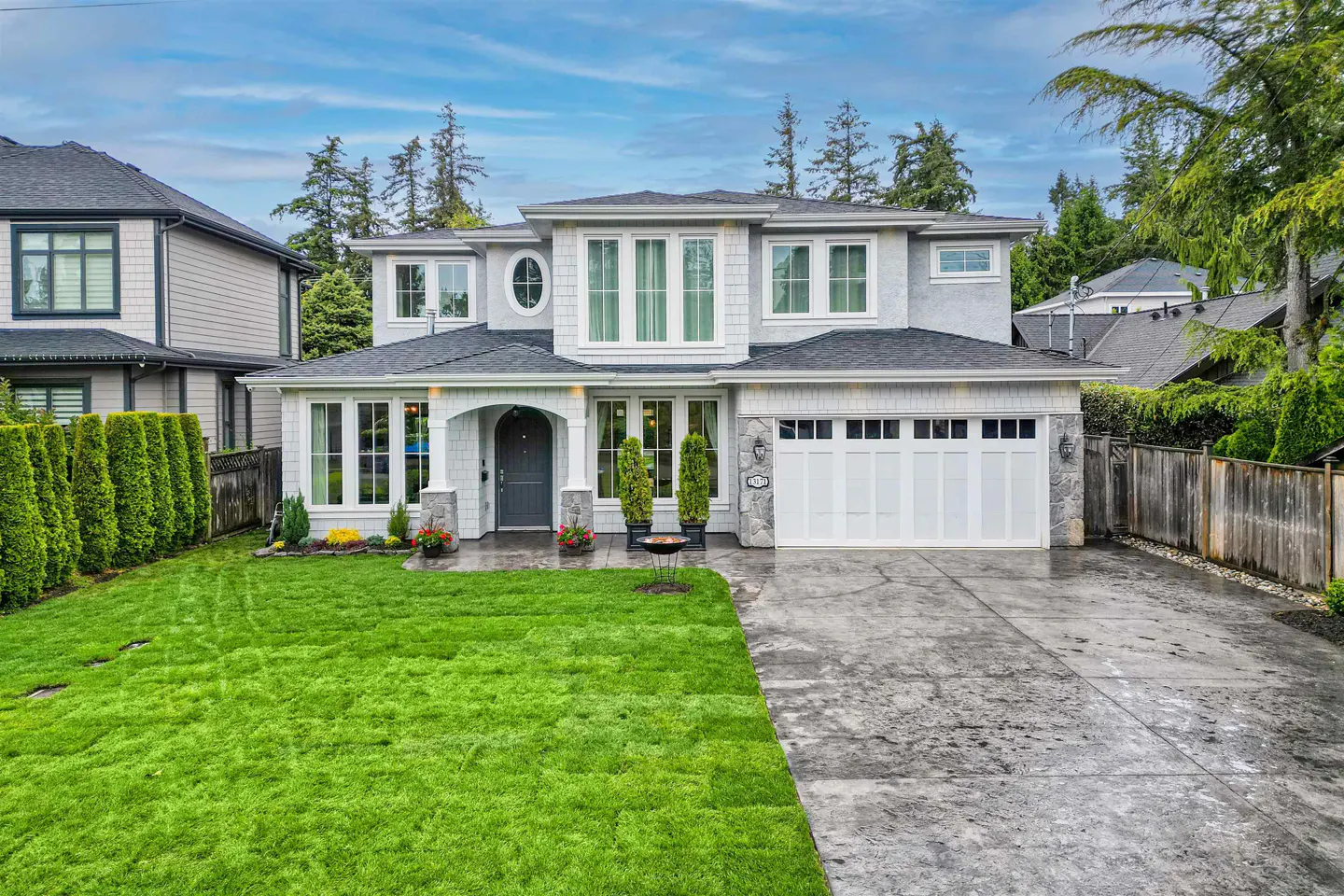 Two-story gray house with white trim, a dark gray front door, and a two-car garage. Green lawn and trees surround the house.