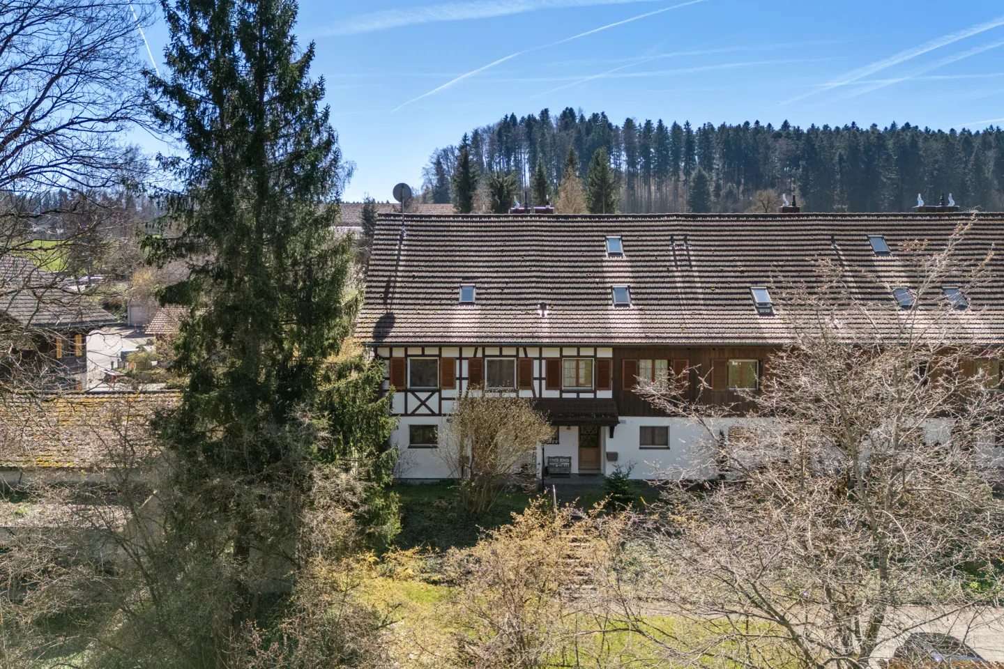 Exterior view of a white, half-timbered house with a brown tiled roof and dormer windows, surrounded by trees.