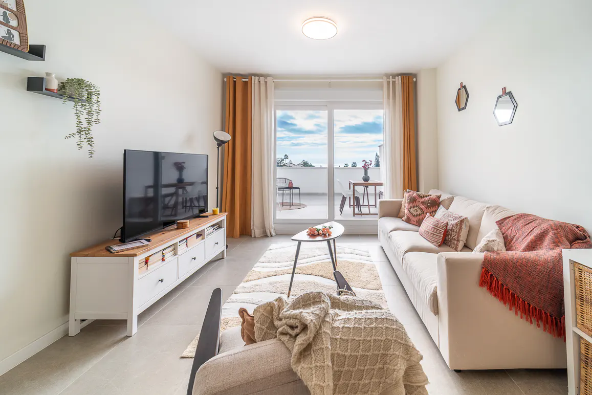 Bright living room with a beige sofa, patterned rug, TV, and balcony with ocean view. Orange curtains frame the sliding glass doors.
