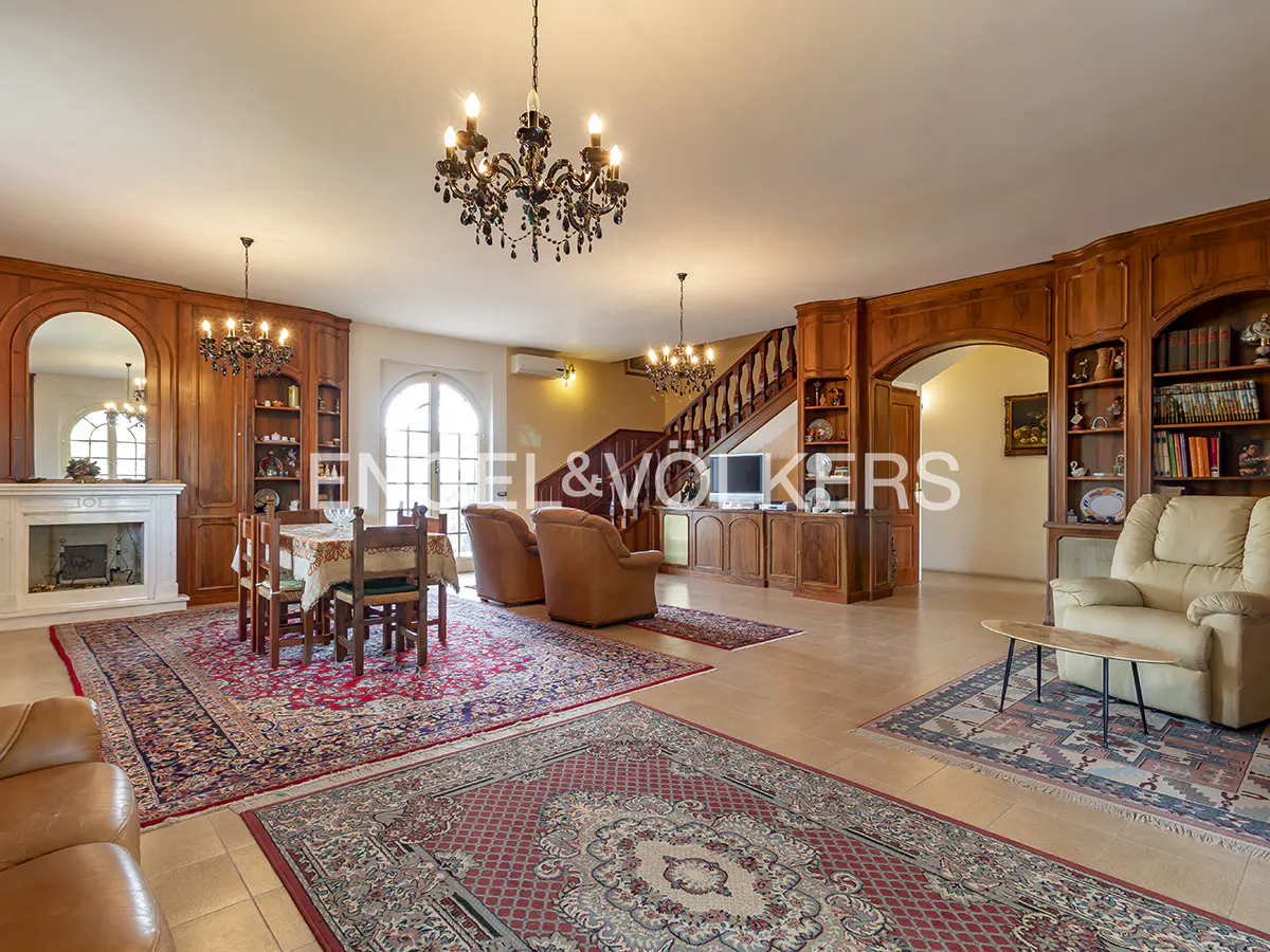 A spacious living room with wood paneling, a fireplace, and a dining table. Two chandeliers hang from the ceiling.