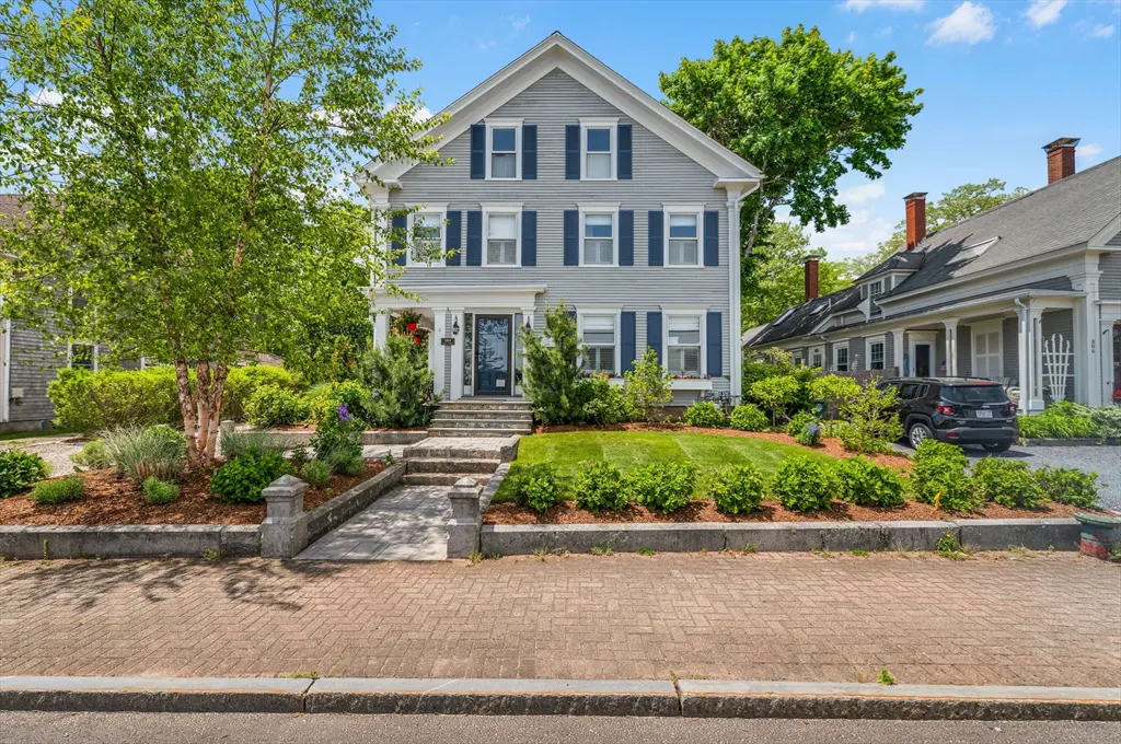 Two-story grey house with blue shutters, a stone walkway, manicured lawn, and a birch tree under a bright blue sky.