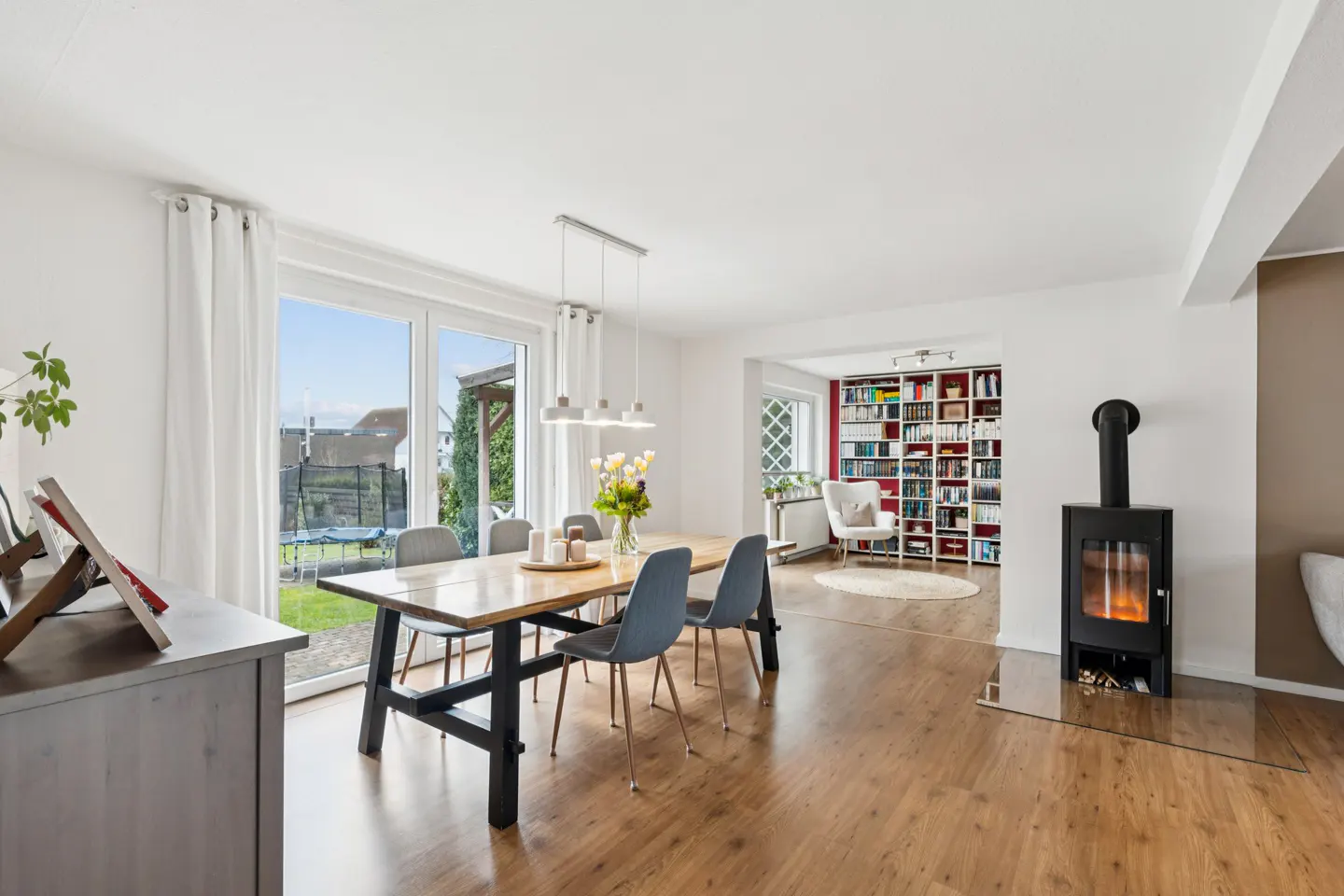 Bright dining room with wood floors, table, and chairs. A fireplace and bookcase are visible in the background. Sliding glass doors lead to a backyard.