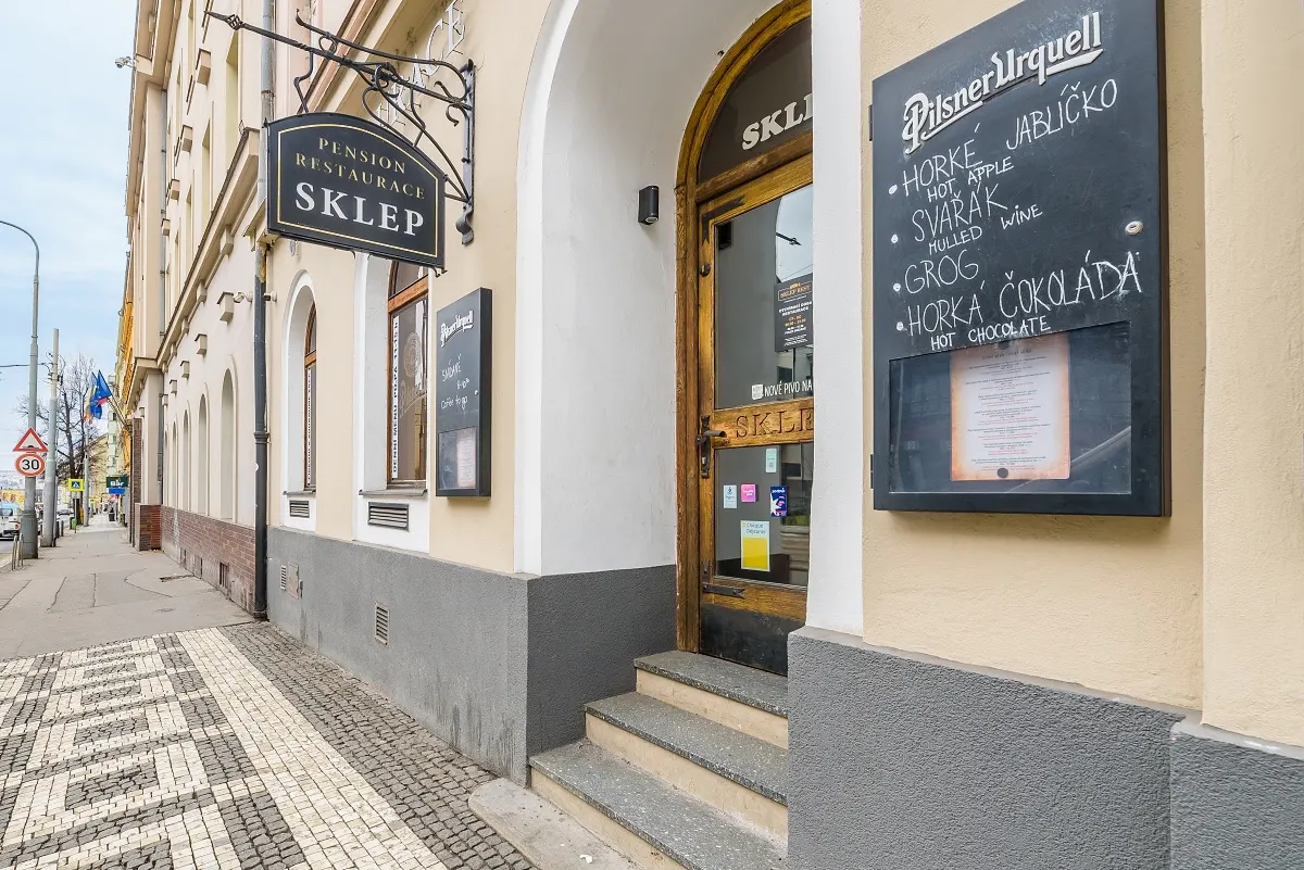 Exterior of Pension Restaurace Sklep with a menu board advertising Pilsner Urquell, hot apple, mulled wine, grog, and hot chocolate.