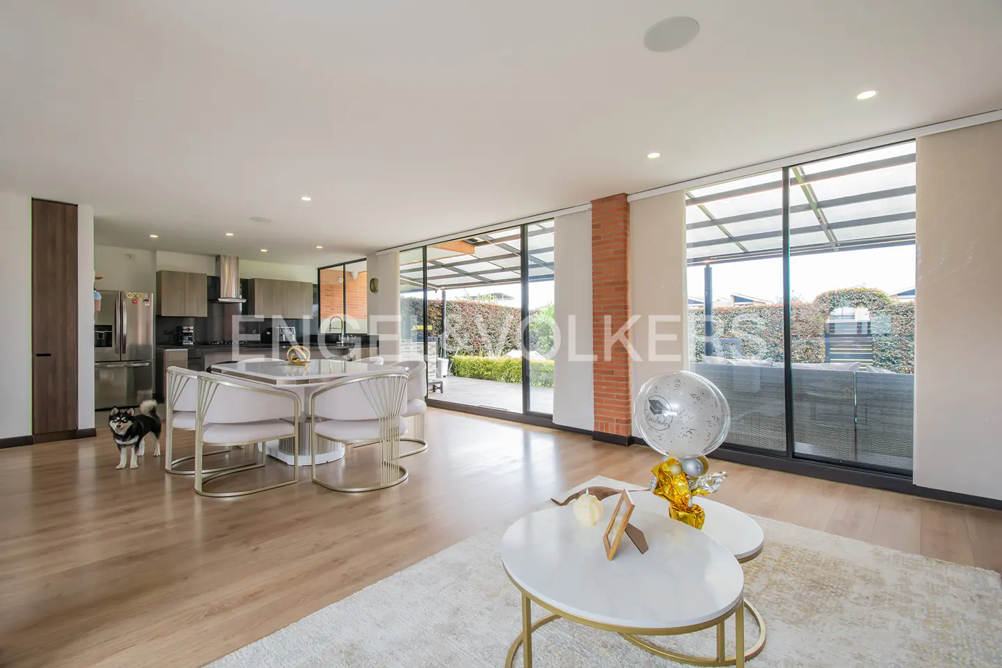 Open-concept living space with wood floors, a dining table with white chairs, and a kitchen in the background. A dog stands near the table.