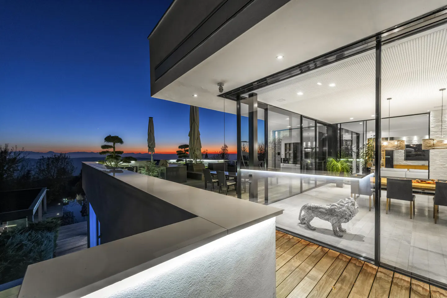 Modern home exterior at dusk with a patio, glass walls, and a lion statue. The sky is blue and orange.